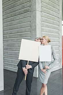 Deutschland, Stuttgart, Geschäftsleute halten leere Schilder in einer Bürohalle und küssen sich - MFPF000245