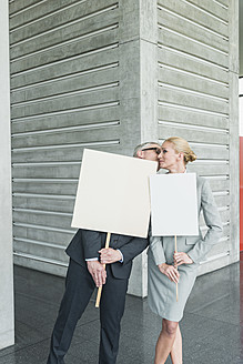 Germany, Stuttgart, Business people holding blank signs in office lobby, kissing - MFPF000245