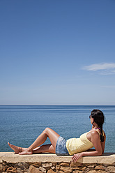 Portugal, mittlere erwachsene Frau, die sich an eine Steinmauer lehnt und auf den Atlantischen Ozean blickt - UMF000357