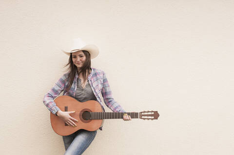 Deutschland, Nordrhein-Westfalen, Junge Frau mit Cowboyhut spielt akustische Gitarre, lächelnd, lizenzfreies Stockfoto