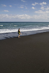 Indonesia, Young woman walking on black sand - MBEF000415