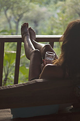 Indonesien, Junge Frau sitzt auf Sofa mit Wasserglas in Veranda - MBEF000441