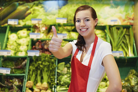 Germany, Cologne, Young woman showing thumbs up in supermarket, smiling, portrait stock photo