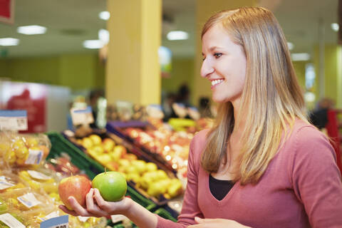 Deutschland, Köln, Junge Frau vergleicht Äpfel im Supermarkt, lizenzfreies Stockfoto