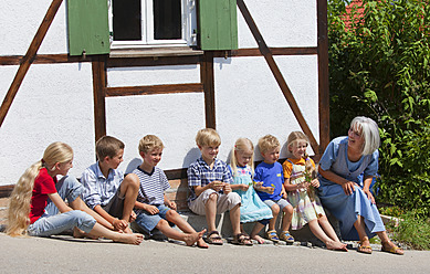Germany, Bavaria, Woman sitting with group of children in front of small house - HSIYF000115