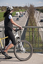 Deutschland, Mittlere erwachsene Frau mit Mountainbike auf Brücke mit Blick auf den Verkehr auf der Autobahn - UMF000513