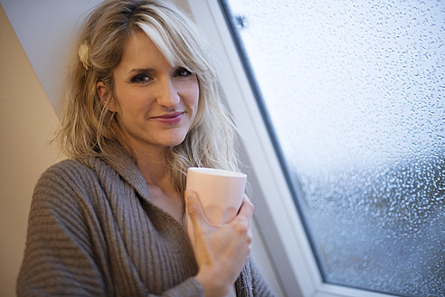 Young woman standing at window with coffee, smiling, portrait - VRF000093