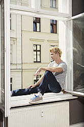 Deutschland, Berlin, Junger Mann sitzt am offenen Fenster - SKF000949