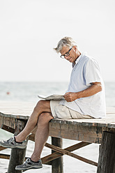 Spain, Senior man reading newspaper on jetty at the sea - JKF000054