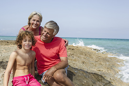 Spanien, Großeltern mit Enkel am Strand sitzend, lächelnd - JKF000095