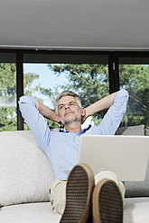 Germany, Berlin, Mature man sitting on sofa using laptop and daydreaming - SKF001160