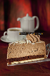 Scheibe Schokoladensahnetorte auf Tortenheber, Kaffeegedeck im Hintergrund - CSF016194