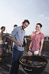 Deutschland, Berlin, Männer und Frauen stehen mit Bierflaschen am Grill, lächelnd - RBF001173