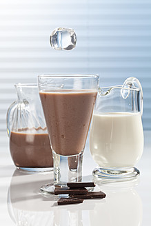 Eiswürfel fallen in ein Glas Schokoladenmilch mit Karaffen und Schokoladenstückchen daneben - CSF016430