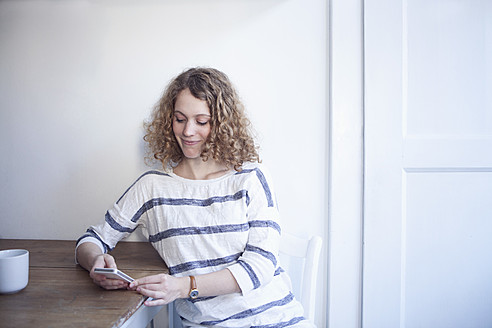 Deutschland, Bayern, München, Junge Frau sitzt am Tisch und benutzt Handy, lächelnd - RBF001115