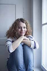 Germany, Bavaria, Munich, Young woman sitting at window, smiling, portrait - RBF001135