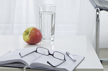 Notizbuch mit Brille, Stift, Apfel und Glas Wasser auf dem Schreibtisch - ASF004826