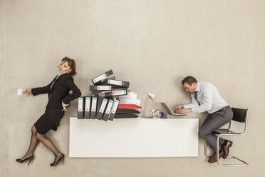 Geschäftsmann arbeitet an einem Schreibtisch, während sich eine Geschäftsfrau auf einen Stapel Akten stützt - BAEF000421