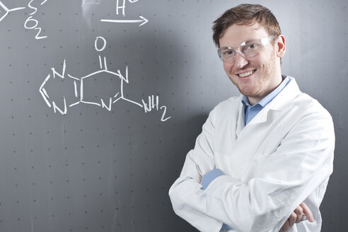 Deutschland, Porträt eines jungen Wissenschaftlers, der neben einer chemischen Gleichung auf einer Kreidetafel steht und lächelt - FLF000298