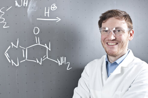 Deutschland, Porträt eines jungen Wissenschaftlers, der neben einer chemischen Gleichung auf einer Kreidetafel steht und lächelt - FLF000288