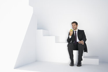 Businessman in black suit sitting on stairs with apple, smiling - PDYF000412