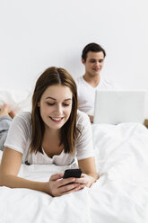 Junge Frau mit Mobiltelefon, während ein junger Mann im Hintergrund einen Laptop benutzt, lächelnd - SPOF000100