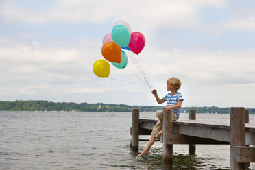 Deutschland, Bayern, Junge sitzt auf Steg und hält bunte Luftballons am Starnberger See - HSIF000247