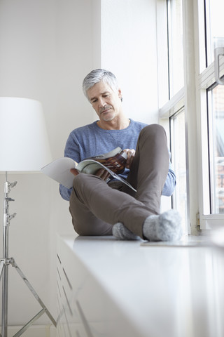 Deutschland, Bayern, München, Älterer Mann sitzt am Fenster und liest Zeitschrift, lizenzfreies Stockfoto