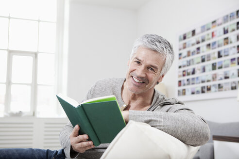 Deutschland, Bayern, München, Porträt eines lächelnden Mannes, der auf der Couch ein Buch liest - RBF001253