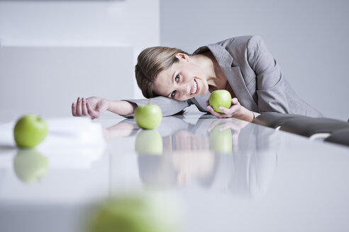 Deutschland, Nordrhein-Westfalen, Köln, Geschäftsfrau auf Konferenztisch liegend mit grünem Apfel in der Hand, lächelnd - FMKF000770