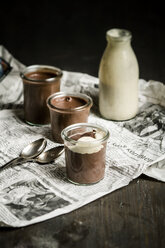 Jars of chocolate pudding with vanilla sauce in bottle on newspaper, close up - SBD000030