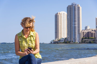 USA, Florida, Miami, Mature woman sitting on surrounding wall of port and using smartphone - ABAF000845
