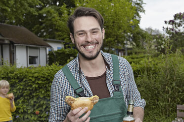 Deutschland, Köln, Porträt eines jungen Mannes mit Wurst und Bierflasche in der Hand, während ein Junge im Hintergrund steht - RHYF000456