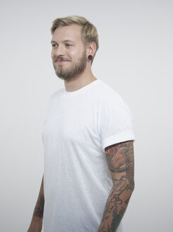Junger Mann mit Tattoos vor weißem Hintergrund, lächelnd - RH000198