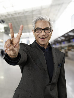 Portrait of businessman showing V sign at airport, smiling - STKF000293