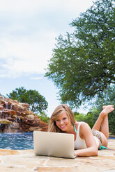Young woman using laptop at swimming pool, smiling - ABAF000879