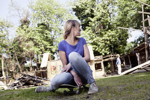 Germany, North Rhine Westphalia, Cologne, Mother sitting on skateboard while son standing in background - FMKYF000504