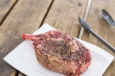 Plate of raw beef bone in rib eye steak with tongs on wooden table, close up - ABAF000957