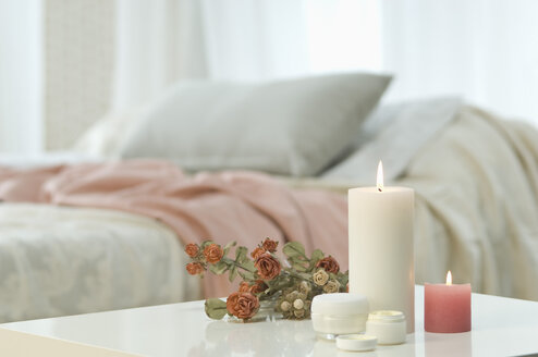 Kerzen, Hautcreme und Rosenstrauß auf Tisch mit Bett im Hintergrund - ASF005038