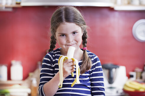 Deutschland, Nordrhein-Westfalen, Köln, Porträt eines Mädchens, das in der Küche eine Banane isst - FMKYF000456