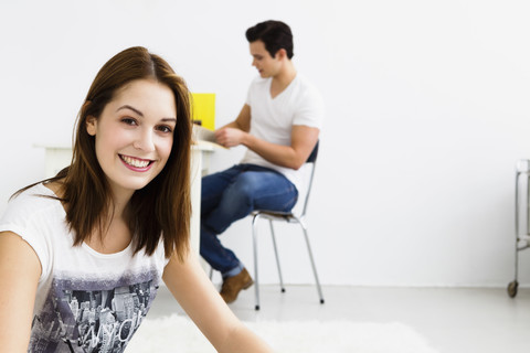 Deutschland, Bayern, München, Porträt einer jungen Frau auf einem Teppich sitzend, im Hintergrund ein junger Mann, lächelnd, lizenzfreies Stockfoto