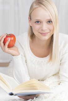 Porträt einer jungen Frau, die auf einer Couch liegt und ein Buch liest, mit einem roten Apfel in der Hand, lächelnd - DRF000123