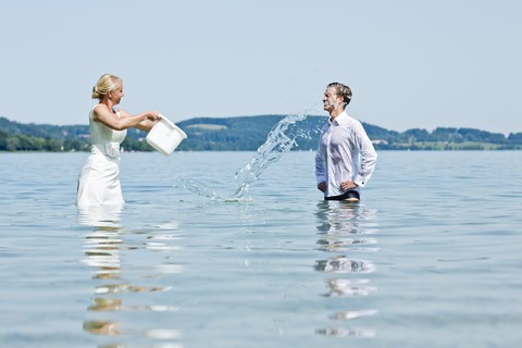 Deutschland, Bayern, Tegernsee, Hochzeitspaar steht im See, gießt Wasser über Bräutigam, lizenzfreies Stockfoto