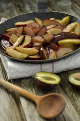 Caramelized prunes in frying pan with wooden spoon and napkin on wooden table, close up - OD000335