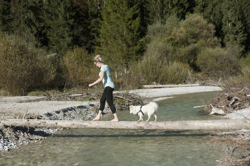 Deutschland, Bayern, Bayerische Alpen, Vorderriss, junge Frau und kleiner Hund balancieren auf einem Baumstamm, um einen Fluss zu überqueren - CRF002481
