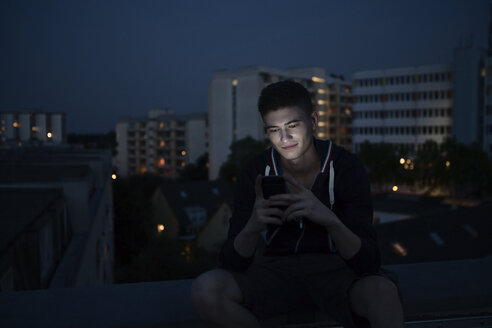 Young man with smartphone sitting on flat roof by night - FEXF000007