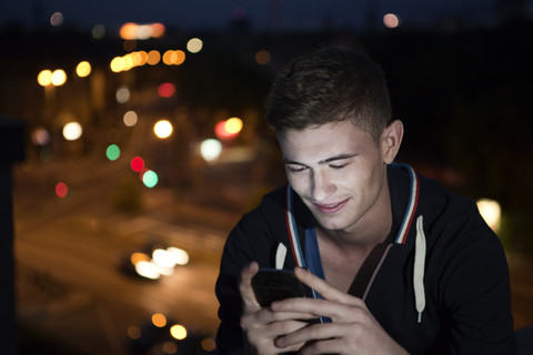 Junger Mann mit Smartphone auf Flachdach sitzend bei Nacht, lizenzfreies Stockfoto