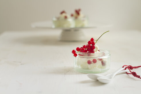 Joghurt-Sahne-Dessert mit roten Johannisbeeren im Glas, zwei weiße Löffel und ein Tortenständer mit Dessertgläsern - SBDF000248