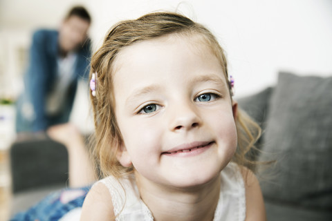 Porträt eines lächelnden jungen Mädchens auf dem Sofa liegend, ihr Vater steht im Hintergrund, lizenzfreies Stockfoto