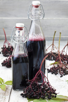 Holunderbeeren (Sambucus) und zwei Flaschen Holundersaft auf weißem Holztisch, Studioaufnahme - CSF020283